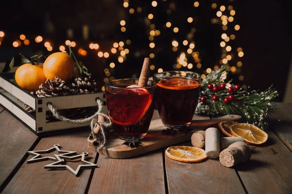 Christmas drinks for the holiday. two glass glasses with mulled wine on a stone board. next to dried oranges, candle and decorative stars, spruce cones. A Christmas tree is burning in the background
