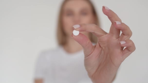A white tablet at arms length in the camera. The girl holds the pill at arms length in focus, then drinks it. Girl on a white background drinking a pill