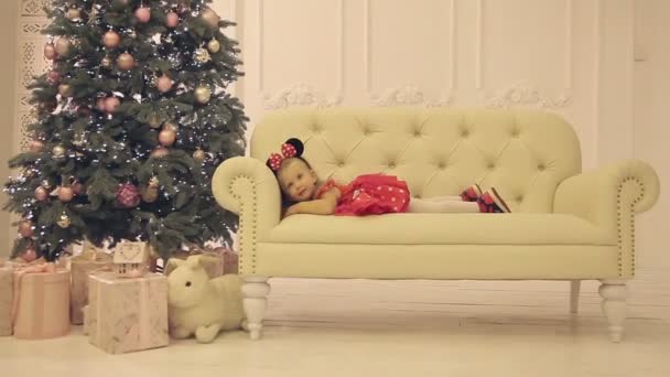 Little cheerful girl sitting on white sofa near the Christmas tree — Stock Video