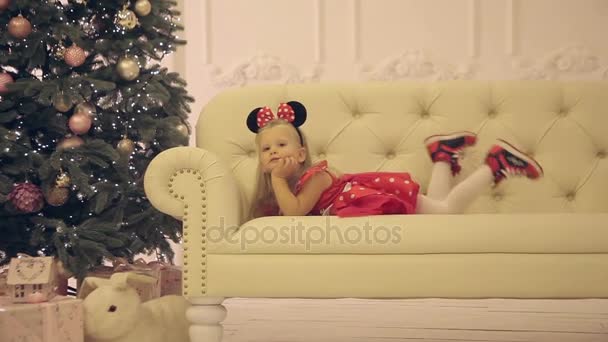 Kleines fröhliches Mädchen sitzt auf weißem Sofa neben dem Weihnachtsbaum — Stockvideo