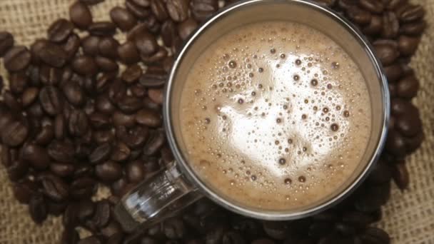Coffee grains around the glass cup with cappuccino on sacking — Stock Video