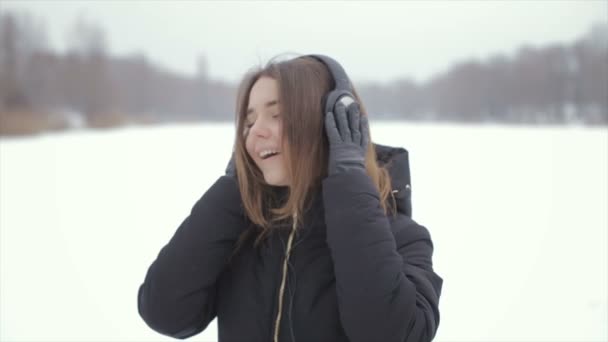 Веселая девушка в зимних наушниках слушает музыку, стоя в снегу — стоковое видео