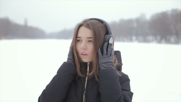 Веселая девушка в зимних наушниках слушает музыку, стоя в снегу — стоковое видео