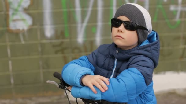 Un niño con gafas de sol se sienta en una bicicleta. Full hd video — Vídeos de Stock
