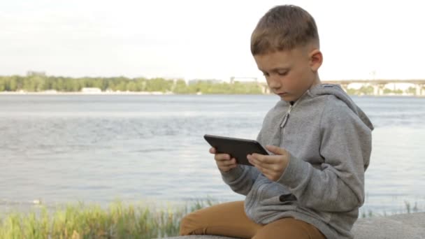 Um menino está brincando em um tablet perto do mar — Vídeo de Stock