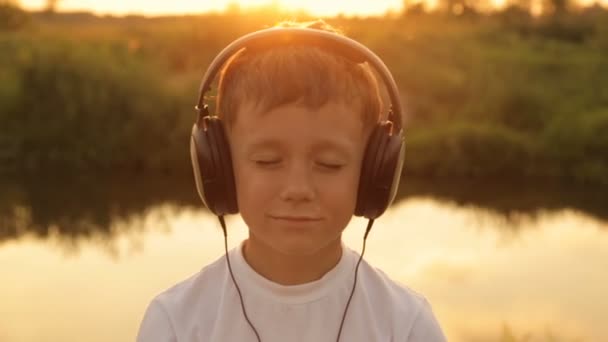 Menino ouvindo música através de fones de ouvido na natureza sob o pôr do sol — Vídeo de Stock