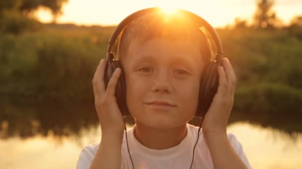 Menino ouvindo música através de fones de ouvido na natureza sob o pôr do sol — Vídeo de Stock