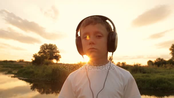 Мальчик слушает музыку через наушники на природе под закатом — стоковое видео