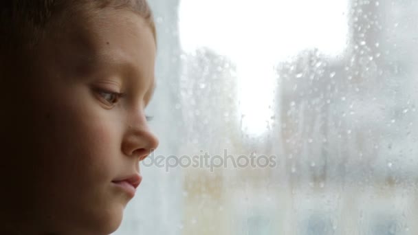 Грустный мальчик смотрит в окно в дождливую погоду — стоковое видео