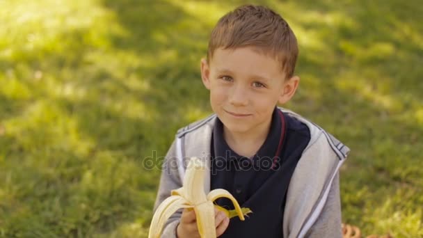 Un niño está comiendo un plátano fresco en un parque de verano — Vídeos de Stock