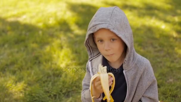 Um menino está comendo uma banana fresca em um parque de verão — Vídeo de Stock