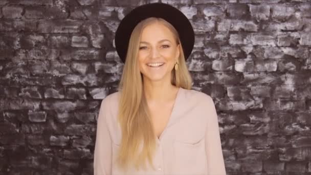 Девушка в черной шляпе и серой рубашке позирует позитивно на фоне текстурной стены — стоковое видео