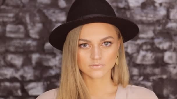 Retrato de una rubia con un sombrero negro sobre un fondo de pared de ladrillo — Vídeos de Stock