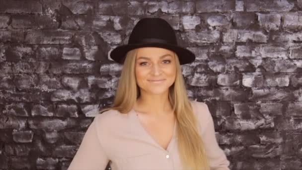 Una chica con un sombrero negro y una camisa gris posando sobre un fondo de pared de ladrillo — Vídeo de stock