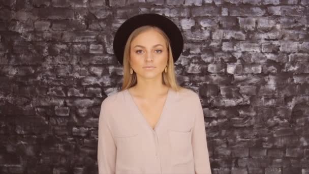 Ένα κορίτσι σε ένα μαύρο καπέλο και ένα γκρι πουκάμισο ποζάρουν με φόντο τοίχο τούβλου — Αρχείο Βίντεο