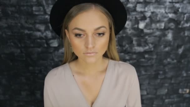 A lány egy fekete kalapot, és egy szürke póló, pózol ellen egy téglafal háttér — Stock videók