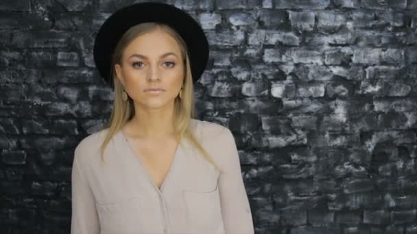 Девушка в черной шляпе и серой рубашке, позирующей на фоне кирпичной стены — стоковое видео