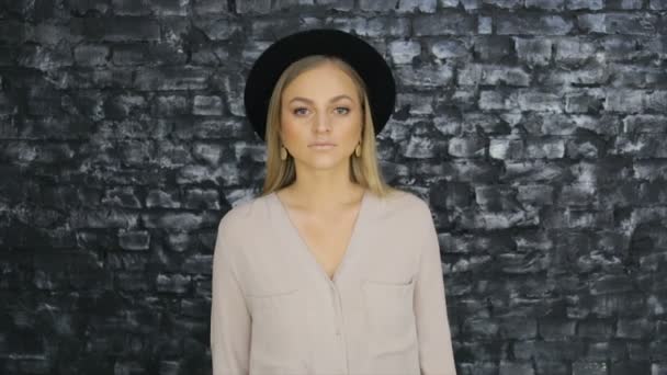 En flicka i en svart hatt och en grå tröja poserar mot tegel vägg bakgrund — Stockvideo