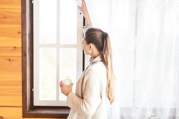 Chica bebe té caliente en la ventana en casa de madera —  Fotos de Stock