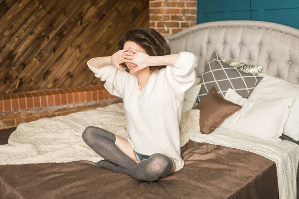 Chica sonriente se sienta en la cama, cerrando los ojos con las manos —  Fotos de Stock