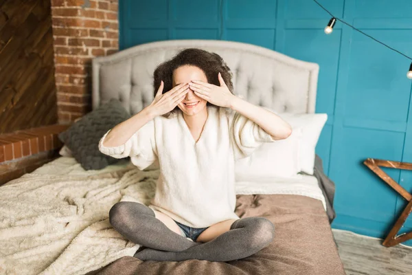 Chica sonriente se sienta en la cama, cerrando los ojos con las manos —  Fotos de Stock