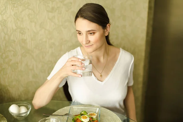 La chica está bebiendo un vaso de agua en la cocina en el tabl —  Fotos de Stock