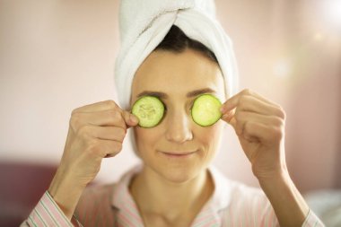 Kafasına havlu geçirmiş bir kadın gözlerine salatalık dilimleri, spa konsepti koymuş.