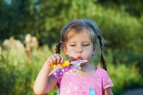La chica en el vestido rosa de 3 años y dos trenzas disfruta de la vida, inflando burbujas. Joy. —  Fotos de Stock