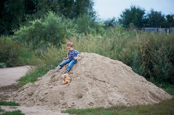 Pequeño niño lindo sentado en una gran pila de arena, jeans elegantes con tirantes y camisa a cuadros. Recuerdos de la infancia sin preocupaciones —  Fotos de Stock