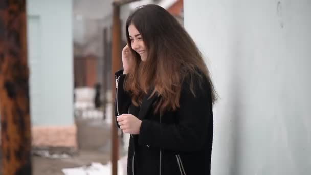 The girl with long hair in a black coat talking on the phone and smiling. On the street. — Stock Video