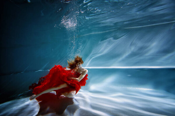 Underwater in the pool with the purest water. Beautiful young girl in a scarlet dress and flowing hair.
