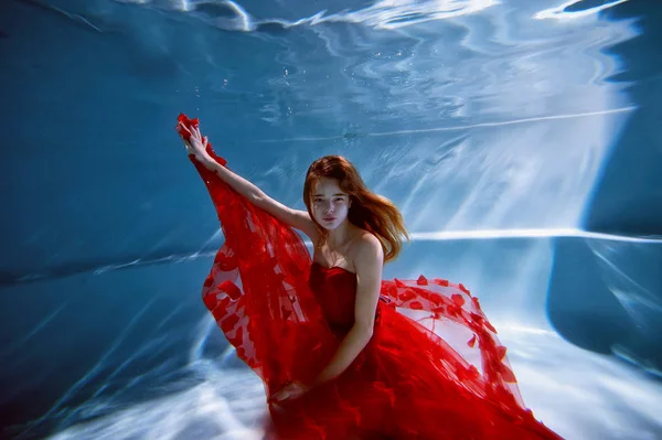 Bajo el agua en la piscina con el agua más pura. Hermosa chica joven en un vestido escarlata y pelo que fluye . —  Fotos de Stock