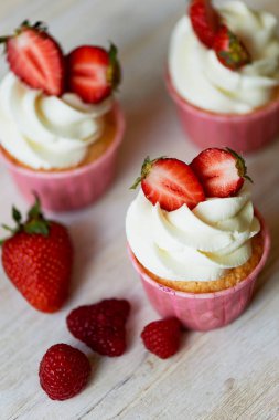 Krem şanti ve tatlı ile cupcakes krem, çilek ile dekore edilmiştir. Bir ışık ahşap arka plan üzerinde.