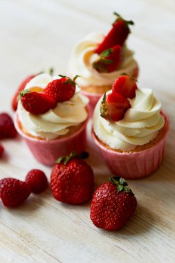 Krem şanti ve tatlı ile cupcakes krem, çilek ile dekore edilmiştir. Bir ışık ahşap arka plan üzerinde.