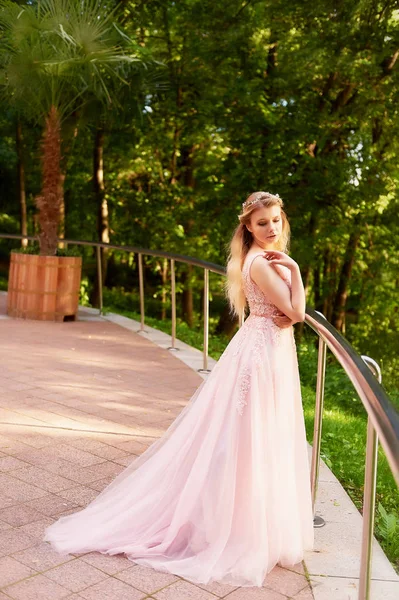 Portret van een meisje van de mooie bruid blond in roze kanten jurk, haar decoratie, handgemaakt. Tederheid. Staande in het midden van een donker bos groen in de stralen van de ondergaande zon. Toont een lange jurk met — Stockfoto