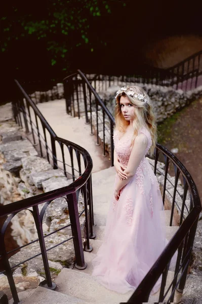 Retrato de una hermosa novia rubia en vestido de encaje rosa, decoración del cabello, hecho a mano. Tiernidad.Es escaleras largas y viejas con piedras, embrague manual —  Fotos de Stock