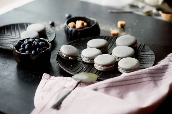 Fotografía monocroma.Pasteles grises macarrones en la superficie oscura de la mesa, junto a una cuchara y un tazón de azúcar con azúcar morena . — Foto de Stock
