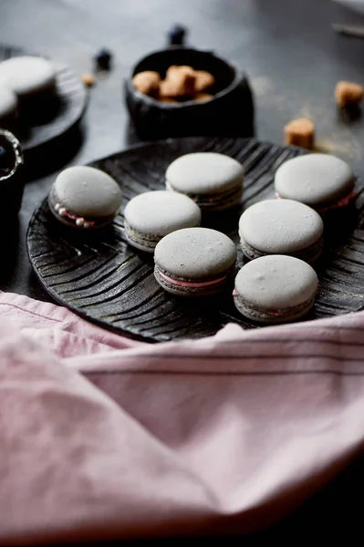 Fotografía monocroma.Pasteles grises macarrones en la superficie oscura de la mesa, junto a una cuchara y un tazón de azúcar con azúcar morena . — Foto de Stock