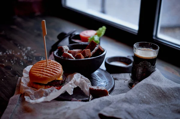 Burger with garlic croutons in the dark interior of the pub. Hearty, quick lunch.Atmospheric photo. Dark beer. — Stock Photo, Image