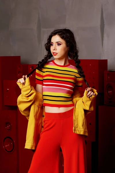 Scatto in studio. La ragazza in un abito luminoso, top e pantaloni svasati nei colori rosso e aranciato.Bruna con i capelli ricci. Mantiene le trecce con le mani — Foto Stock