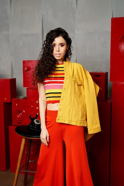 Scatto in studio. La ragazza in un abito luminoso, top e pantaloni svasati nei toni del rosso e dell'arancio. Bruna con i capelli ricci. Posizione bella e insolita. Attraente — Foto Stock