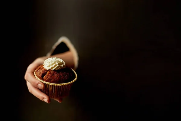 Um buraco no papel, um buraco. Mão segurando um cupcake de chocolate com nata.Confeitaria, dieta. Espaço de cópia, tendência. O fundo é preto . — Fotografia de Stock