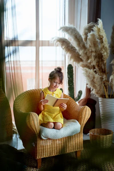 Niña, 6 años. Le tiraron del pelo en un bollo. Lee el libro con placer. Vestido amarillo, hermoso interior.Anhelo de aprender, amor por la lectura . —  Fotos de Stock