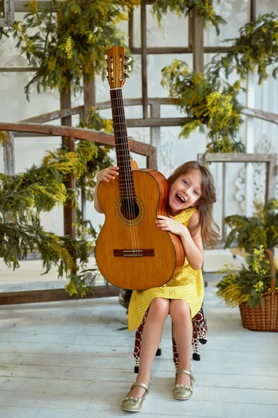 Niña, 6 años. Tiene el pelo suelto. Vestido amarillo, Mimosa, hermoso interior. Riendo y sosteniendo una guitarra. Clase de música . —  Fotos de Stock