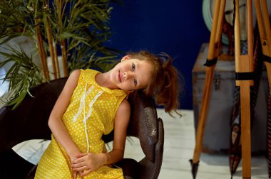 Sarı elbiseli gülümseyen bir kızın portresi. 6 yaşında. Kulağı delinmiş. Mavi gözlü, uzun saçlı..