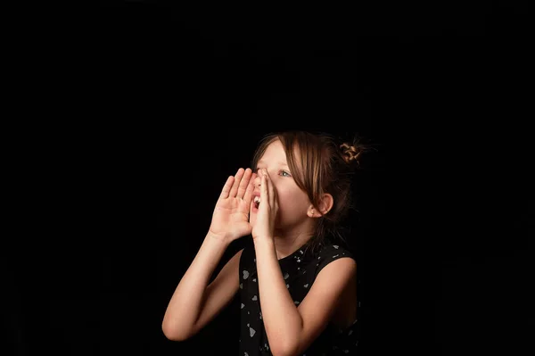A menina tem 6 anos, gritando, mãos no rosto na forma de uma boca.Para gritar.Uma menina de 6 anos, em um fundo preto . — Fotografia de Stock