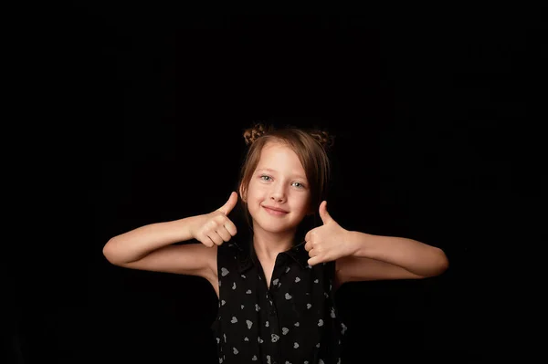 Una niña de 6 años, sobre un fondo negro.Muestra gesto de clase, todo va a estar bien. Dedos arriba . — Foto de Stock