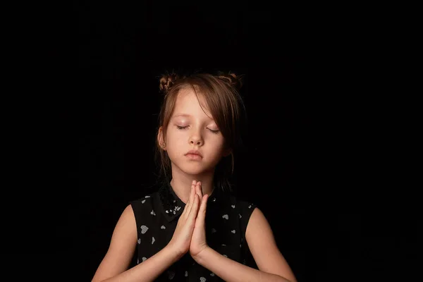 Uma menina de 6 anos, contra um fundo escuro. Um gesto de oração e gratidade.As palmas das mãos estão dobradas, os olhos fechados . — Fotografia de Stock