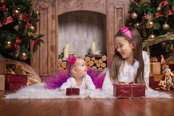 Söta systrar liggande på vit matta nära julgranar, klädd i rosa kjolar och röda hårband. Ler småbarn. Julklappar. — Stockfoto
