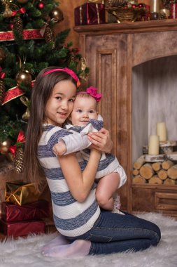 Noel ağacı ve şömine yakın beyaz halı üzerinde oturan sevimli kız giyiyor kazak ve kırmızı saç bantları çizgili. Gülümseyen küçük çocuklar. Noel hediyeleri.